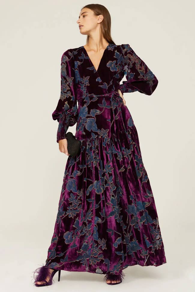 Model wearing purple and navy blue floral velvet maxi dress with V-neckline and balloon sleeves paired with feather purple heels on a white background