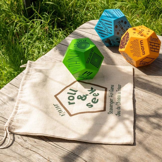 Three geometric dice with enactment   prompts connected  a woody  table, adjacent  to a cloth container  labeled 