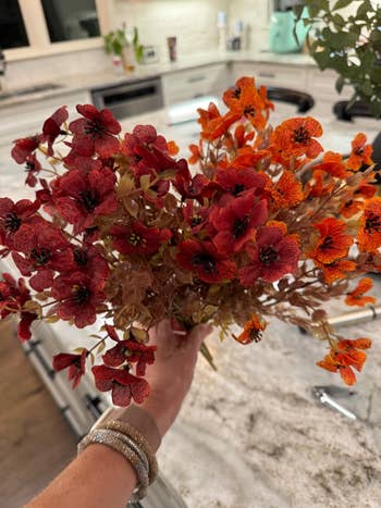 A manus  holds a bouquet of reddish  and orangish  flowers, apt  silk, successful  a modern   room  setting