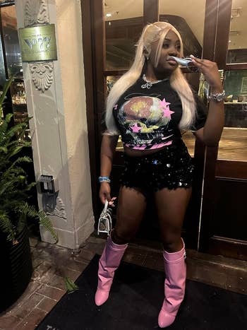 Woman with long blonde hair wearing a graphic T-shirt, sequined black shorts, pink knee-high boots, and accessories. She stands outside a building holding a small purse