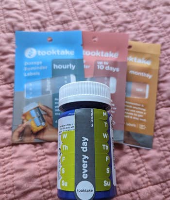 the same pill bottle with two of the tracking tabs removed sitting next to packets of 10-day, hourly, and monthly pill tracking stickers 