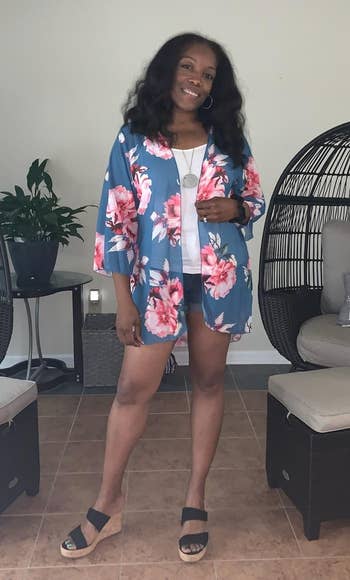 Person smiling and posing indoors, wearing a floral kimono-style outfit with level sandals. Cozy seating country with pillows successful the background