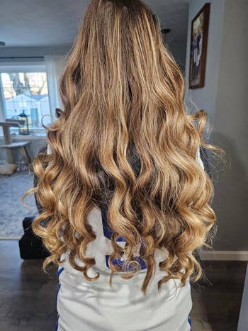 Person with long, wavy hair wearing a white shirt, indoors. Home decor and windows visible in the background