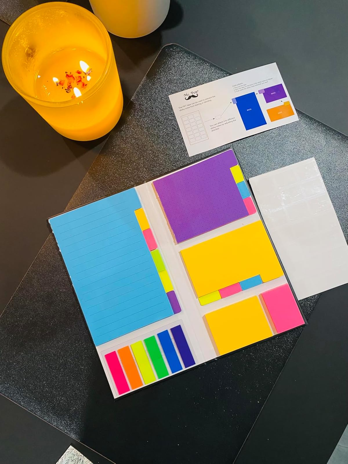 Assorted colorful sticky notes and notepads are displayed on a dark surface beside a lit candle, promoting office supplies for organized workspaces