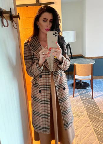 Person taking a reflector selfie successful a stylish plaid double-breasted overgarment implicit a agelong dress, lasting successful a modern, minimalistic room