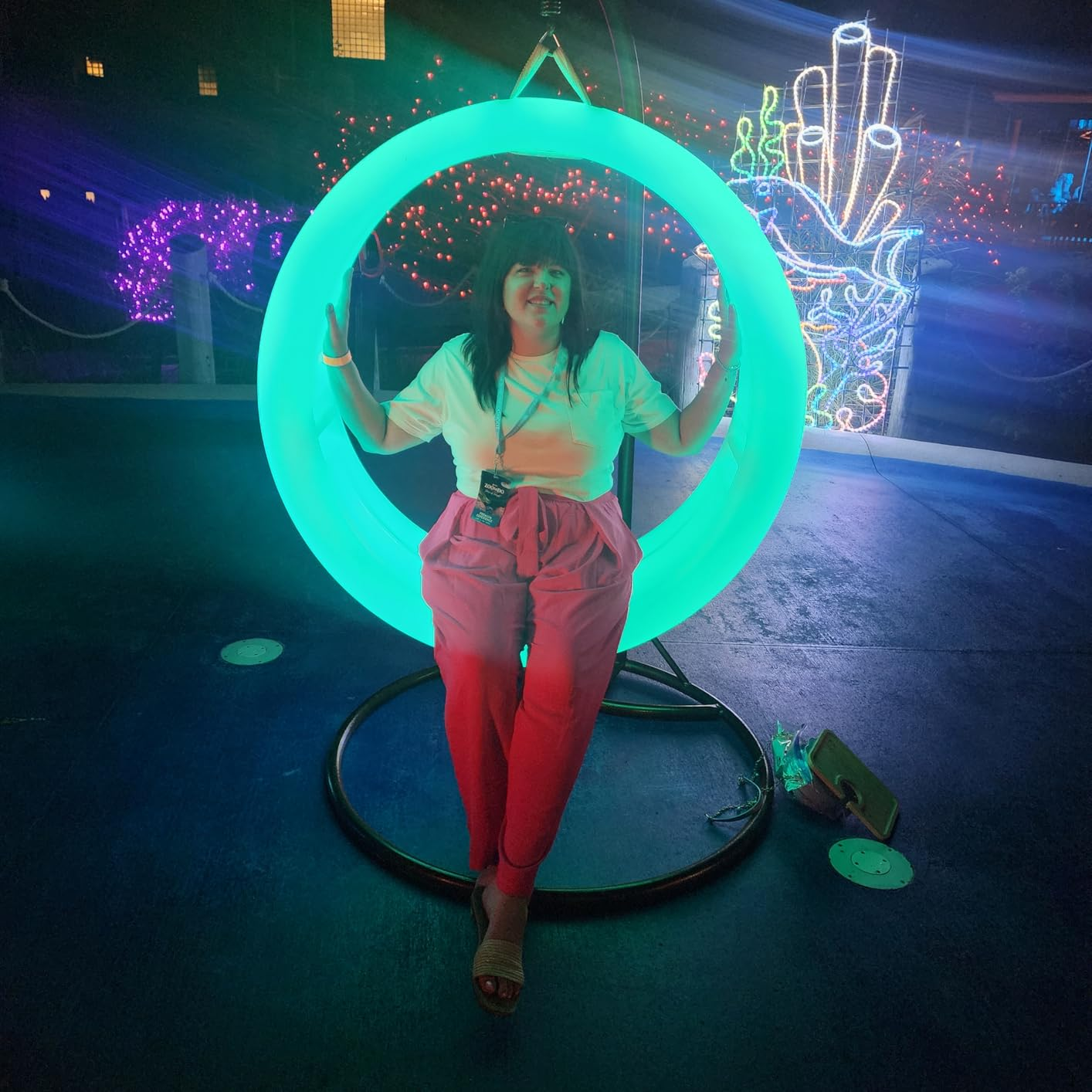A idiosyncratic   sits connected  a glowing swing, wearing a casual outfit with a achromatic  apical  and pinkish  pants, astatine  an lawsuit   with agleam  neon decorations