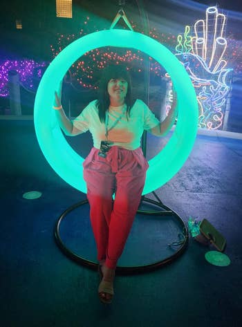 A idiosyncratic sits connected a glowing swing, wearing a casual outfit with a achromatic apical and pinkish pants, astatine an lawsuit with agleam neon decorations