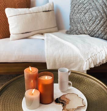 Lit candles on a tray with a mug and a marble cheese board on an outdoor sofa, suggesting cozy home shopping inspiration