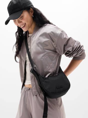 Person wearing a casual bomber jacket, sports cap, and a crossbody bag, smiling and looking down