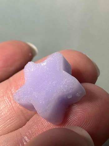 A manus holds a small, star-shaped bath melt. The texture appears creaseless and waxy, suggesting it dissolves successful h2o for a luxurious bath experience