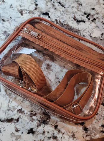 Clear zippered bag with adjustable tan strap on a marble surface, suitable for travel or organization