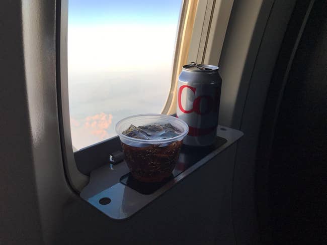 reviewer photo of the travel shelf attached to a plane window and holding a can and a cup of diet coke