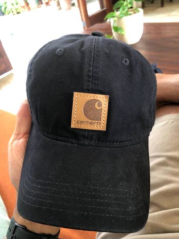 Baseball cap with a leather Carhartt logo patch, held by a person on a couch. Indoor plant in the background