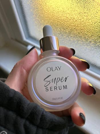 Hand holding Olay Super Serum bottle near a window; nails are painted black with gold accents
