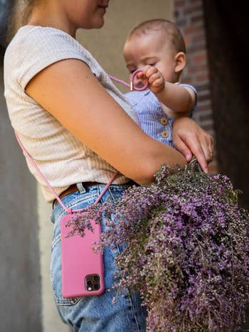 Model holding a baby with their phone in a pink crossbody case strapped across their body 
