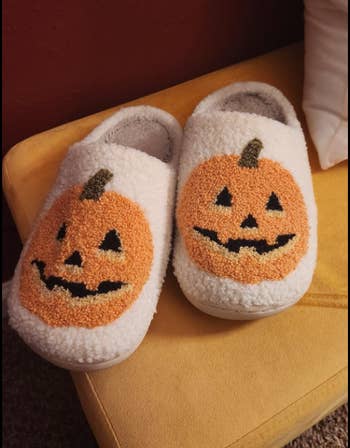 Soft slippers with pumpkin faces design, placed connected a chair, promoting cozy and festive Halloween-themed footwear