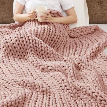 A person sitting on a bed with a chunky knit blanket and holding a mug. The image emphasizes coziness and comfort