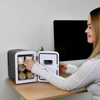 Person placing beverages and a snack cup into a compact mini fridge on a desk, next to a computer