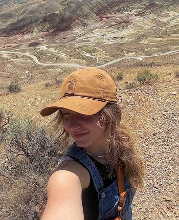 Person in overalls and a cap takes a selfie on a mountain trail, with hills and sky in the background
