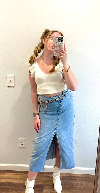 reviewer in mirror selfie wearing a cream top, denim skirt, white boots, and a chain belt