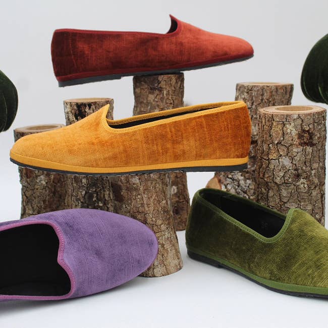 Four velvet loafers in red, mustard, purple, and green displayed with wood logs