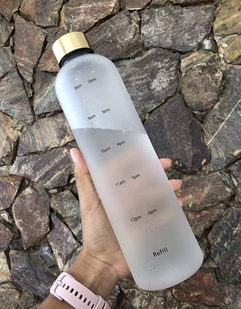 reviewer holding clear frosted water bottle with gold cap and time marks in black
