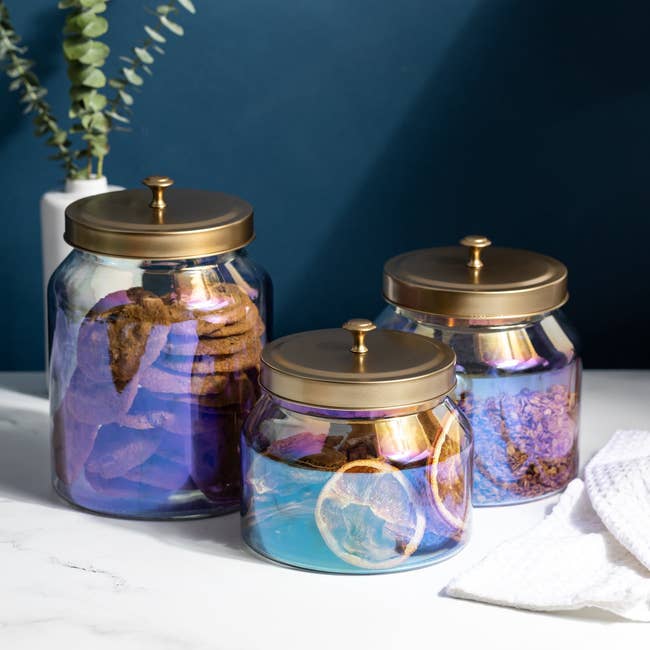 Three solid jars with aureate lids containing cookies and dried citrus slices, placed connected a marble surface, with a works successful the background