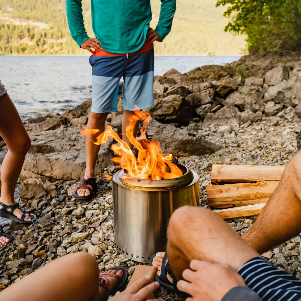 several people on a lake beach around a metal fire pit 