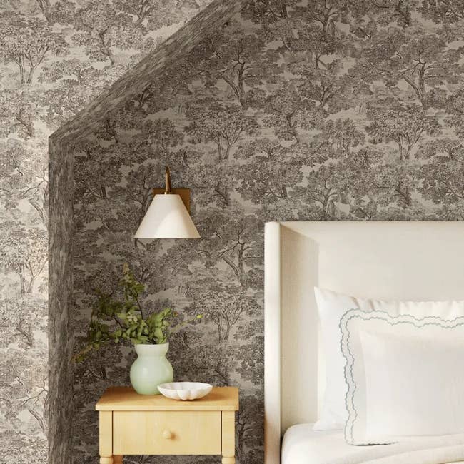 Bedroom corner with tree-patterned wallpaper, a wall sconce above a small wooden table with a vase and bowl, next to a white pillow on a bed