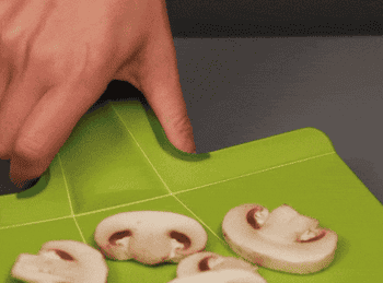 gif of someone folding up the green cutting board with sliced mushrooms on it and pouring the mushrooms into a pan