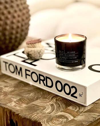 A lit soy candle is placed on a Tom Ford 002 book on a rustic wooden table, with decorative items around it