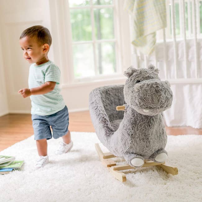 Toddler next to a plush hippo rocker in a bright room, smiling, invokes a playful mood for shopping