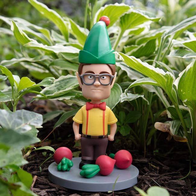 Dwight schrute figure with glasses in a garden setting, wearing a green pointy hat, yellow shirt, red bow tie, and suspenders, surrounded by vegetables