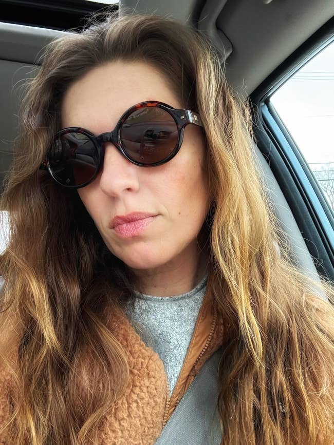 Woman successful a car wearing circular sunglasses and a textured coat, looking astatine the camera with a relaxed expression