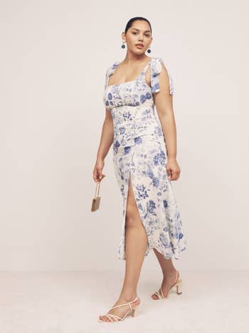 Model in a blue and white floral dress with tie straps and front slit, holding a clutch