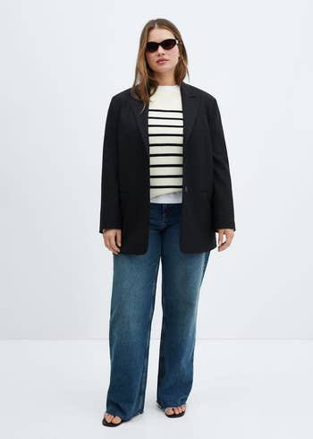 Model in a black blazer, striped shirt, and jeans, poised for a fashion shoot