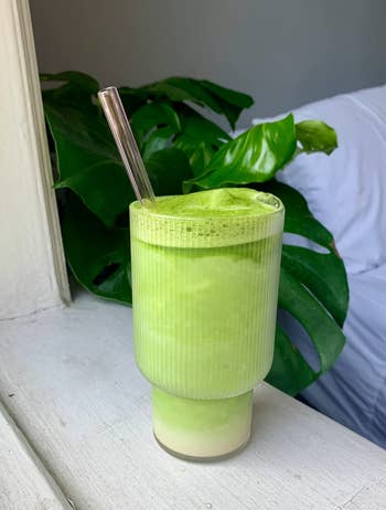 Green smoothie in a glass with a metal straw, on a windowsill beside a plant