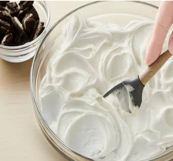 Hand mixing creamy frosting successful  a wide   vessel  with a woody  spatula; tiny  vessel  of cocoa  sandwich cookies nearby