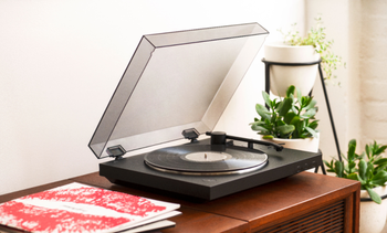 Black turntable with black needle and clear cover on top of wooden accent table with stacked vinyls