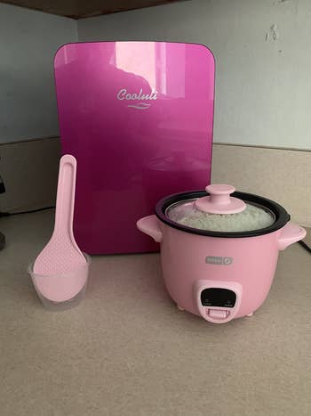 a reviewer photo of the mini rice cooker in pink on a counter in front of a mini fridge