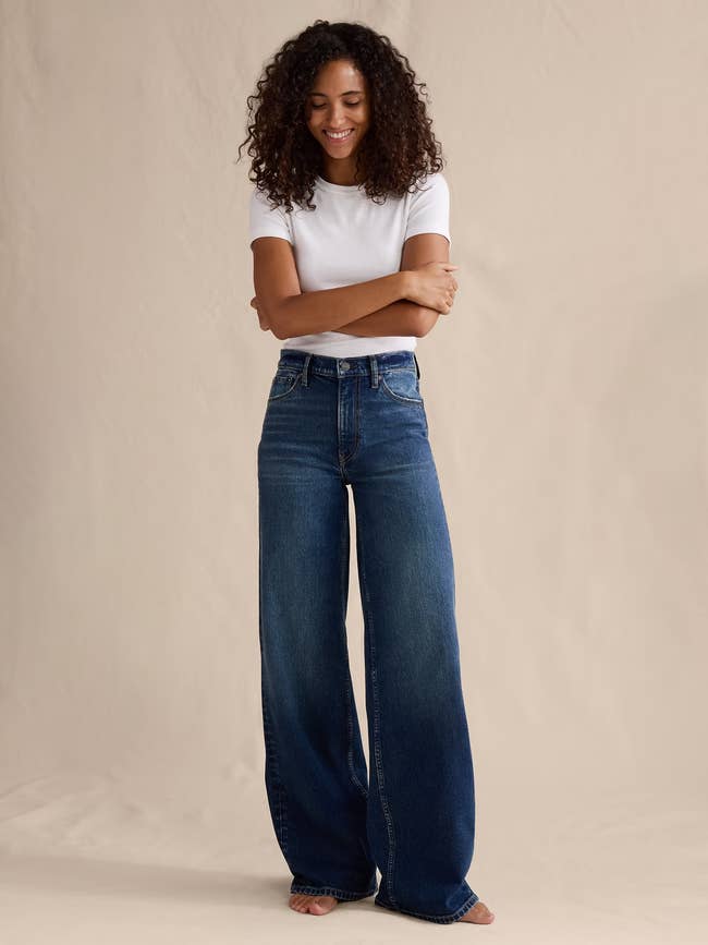 Person successful achromatic t-shirt and wide-leg jeans stands barefoot with arms crossed, smiling, against a plain background