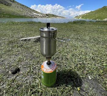 Portable stove with a pot set on grass with a lake and mountains in the background, emphasizing outdoor cooking equipment