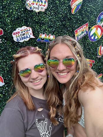 Two people smiling, wearing reflective sunglasses and casual tops, stand in front of a greenery wall decorated with peace and pride-themed signs