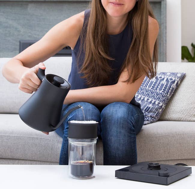 person pouring hot beverage into glass from black tea kettle while sitting on couch