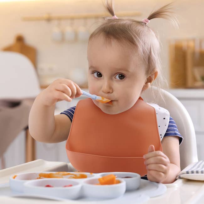 A toddler successful a precocious seat eats with a spoon from a divided plate. The kid wears a bib with a ponytail style. Kitchen mounting successful the background