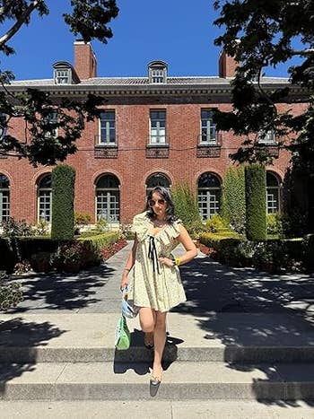 Person stands on steps wearing a short, ruffled dress with black accents, holding a green and white bag, in front of a brick building