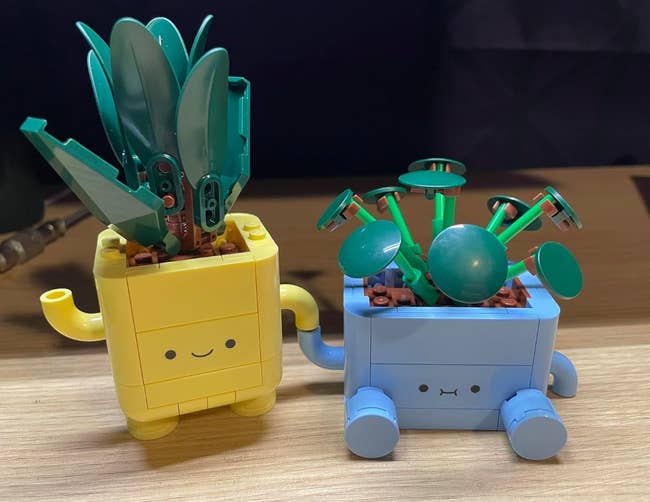 Two lego planter figures with cute faces, one with green leaf pieces and another with round green pieces, on a wooden surface