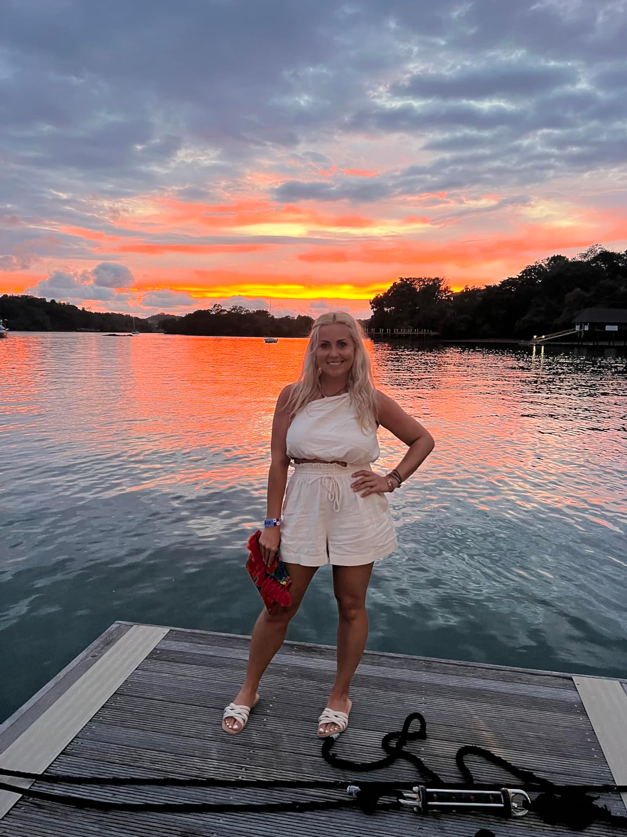 Reviewer in a sleeveless romper holding a red clutch stands on a dock at sunset over calm water