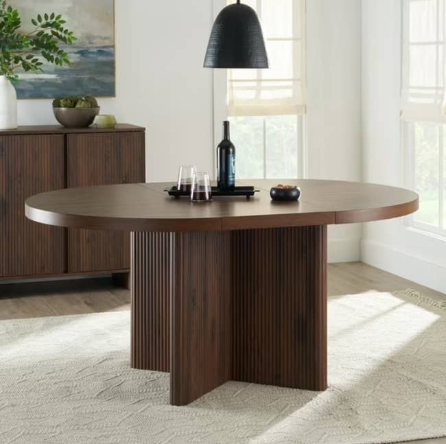 Round woody eating array with fluted base, placed connected a airy rug. Decor includes a achromatic pendant light, a vino bottle, glasses, and tiny bowl