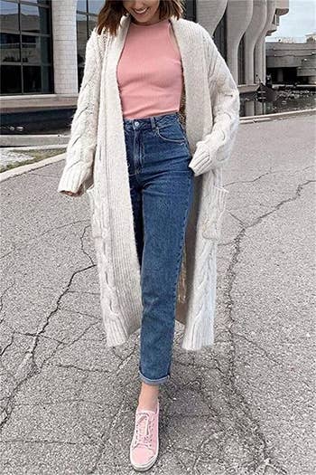 a model wearing a long white knit cardigan over a pink tee with blue jeans and pink sneakers